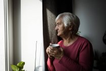 Задумчивая пожилая женщина пьет чай, глядя в окно — стоковое фото