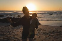 Mãe carregando seu filho na praia durante o pôr do sol — Fotografia de Stock