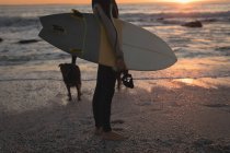 Surfista con tavola da surf in piedi sulla spiaggia al tramonto — Foto stock