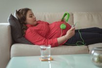 Jovem grávida deitada no sofá mulher colocando fones de ouvido em sua barriga em casa — Fotografia de Stock