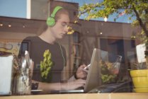 Чоловік слухає музику на навушниках під час використання мобільного телефону та ноутбука в кафе — стокове фото
