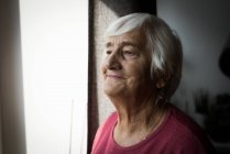 Задумчивая пожилая женщина, стоящая у окна дома — стоковое фото