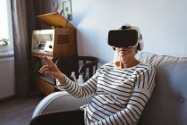 Donna anziana che indossa un auricolare realtà virtuale mentre si rilassa su un divano — Foto stock