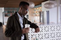 Бизнесмен смотрит на умные часы, выпивая кофе в кафетерии в креативном офисе — стоковое фото