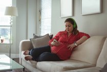 Jeune femme enceinte assise sur un canapé listant de la musique sur son téléphone portable à la maison — Photo de stock