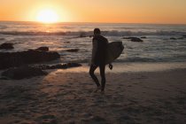Surfista caminando con tabla de surf en la playa al atardecer - foto de stock
