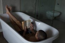 Junger Mann liest Buch, während er in Badewanne im Badezimmer liegt — Stockfoto