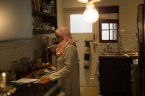 Mulher muçulmana falando ao telefone e usando laptop na cozinha — Fotografia de Stock