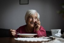 Усміхаючись старший жінка говорити на мобільний телефон, маючи сніданок у вітальні — стокове фото
