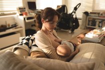 Молодая мама сидит на диване и кормит грудью своего ребенка в гостиной дома — стоковое фото