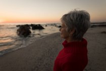 Femme âgée réfléchie debout sur la plage pendant le coucher du soleil — Photo de stock