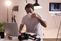 Fotógrafo masculino tomando café enquanto usa laptop no estúdio de fotos — Fotografia de Stock