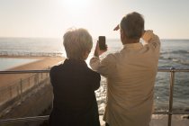 Vista trasera de pareja mayor usando teléfono móvil cerca del lado del mar en un día soleado - foto de stock