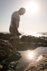Вид сбоку на пожилого человека, стоящего на камне у моря в солнечный день — стоковое фото