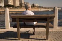 Vista trasera de la mujer mayor sentada en el banco junto al mar en el paseo marítimo - foto de stock