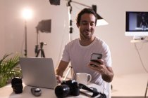 Чоловічий фотограф використовує мобільний телефон під час роботи на ноутбуці у фотостудії — стокове фото