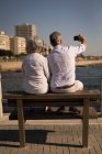 Vista trasera de la pareja mayor tomando selfie mientras está sentada en el banco en el paseo marítimo - foto de stock