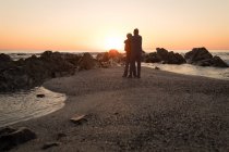 Вид спереду старшої пари, що обіймається на пляжі під час заходу сонця — стокове фото