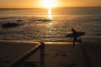 Surfista com prancha de surf correndo na praia durante o pôr do sol — Fotografia de Stock
