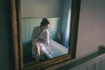 Рефлексия женщины, сидящей на ванне и проверяющей воду в ванной дома — стоковое фото