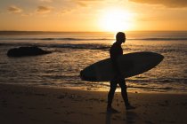 Surfista con tabla de surf caminando por la playa al atardecer - foto de stock