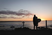 Вид сзади на человека с доской для серфинга, стоящей на пляже во время заката — стоковое фото