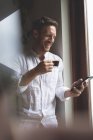 Щасливий чоловік використовує мобільний телефон, маючи каву вдома — стокове фото