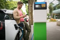Homme utilisant un téléphone portable tout en rechargeant la voiture électrique à la station de charge — Photo de stock