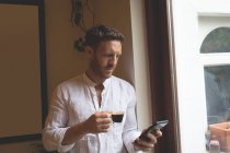 Homem usando telefone celular enquanto toma café preto em casa — Fotografia de Stock