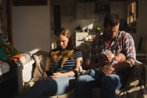 Mother using mobile phone while father feeding his baby on sofa at home — Stock Photo
