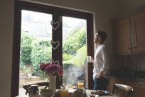 Hombre reflexivo tomando café mientras está de pie cerca de la ventana en casa - foto de stock