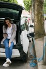 Woman using mobile phone while charging electric car at charging station — Stock Photo