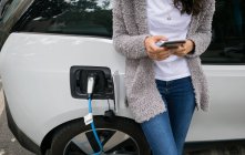 Partie médiane de la femme utilisant un téléphone portable tout en rechargeant la voiture électrique à la station de charge — Photo de stock