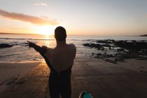 Vista traseira do surfista vestindo traje na praia durante o pôr do sol — Fotografia de Stock