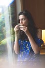 Bella donna che prende il caffè mentre in piedi vicino alla finestra a casa — Foto stock