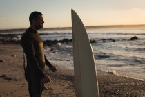 Surfista con tavola da surf in piedi sulla spiaggia durante il tramonto — Foto stock