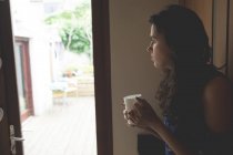 Задумчивая женщина пьет кофе, стоя у окна дома — стоковое фото