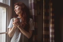 Задумчивая женщина с кофейной чашкой, стоящей у окна дома — стоковое фото