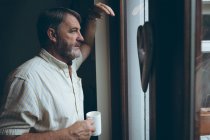 Homme âgé réfléchi avec tasse de café regardant par la fenêtre à la maison — Photo de stock