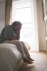 Задумчивая пожилая женщина сидит на кровати в спальне дома — стоковое фото
