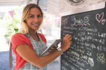 Portrait of female waitress writing menu on menu board while using digital tablet — Stock Photo