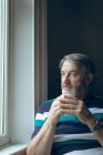 Продуманий старший чоловік дивиться крізь вікно, маючи каву вдома — стокове фото
