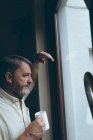 Задумчивый пожилой человек с кофейной чашкой, смотрящий в окно дома — стоковое фото