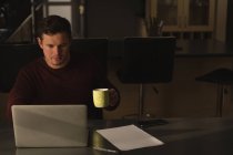 Mann trinkt Kaffee, während er Laptop auf Esstisch zu Hause benutzt — Stockfoto
