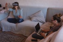 Couple lesbien utilisant un casque de réalité virtuelle et un ordinateur portable sur le canapé à la maison — Photo de stock