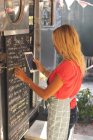 Beautiful female waitress writing menu on menu board while using digital tablet — Stock Photo