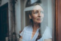 Femme âgée réfléchie regardant par la fenêtre à la maison — Photo de stock