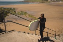 Surfista con tabla de surf caminando en la escalera cerca de la playa - foto de stock