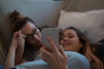 Pareja lesbiana usando teléfono móvil en el sofá en casa - foto de stock