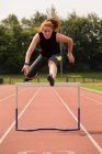 Young female athletic running over hurdle on sports track — Stock Photo